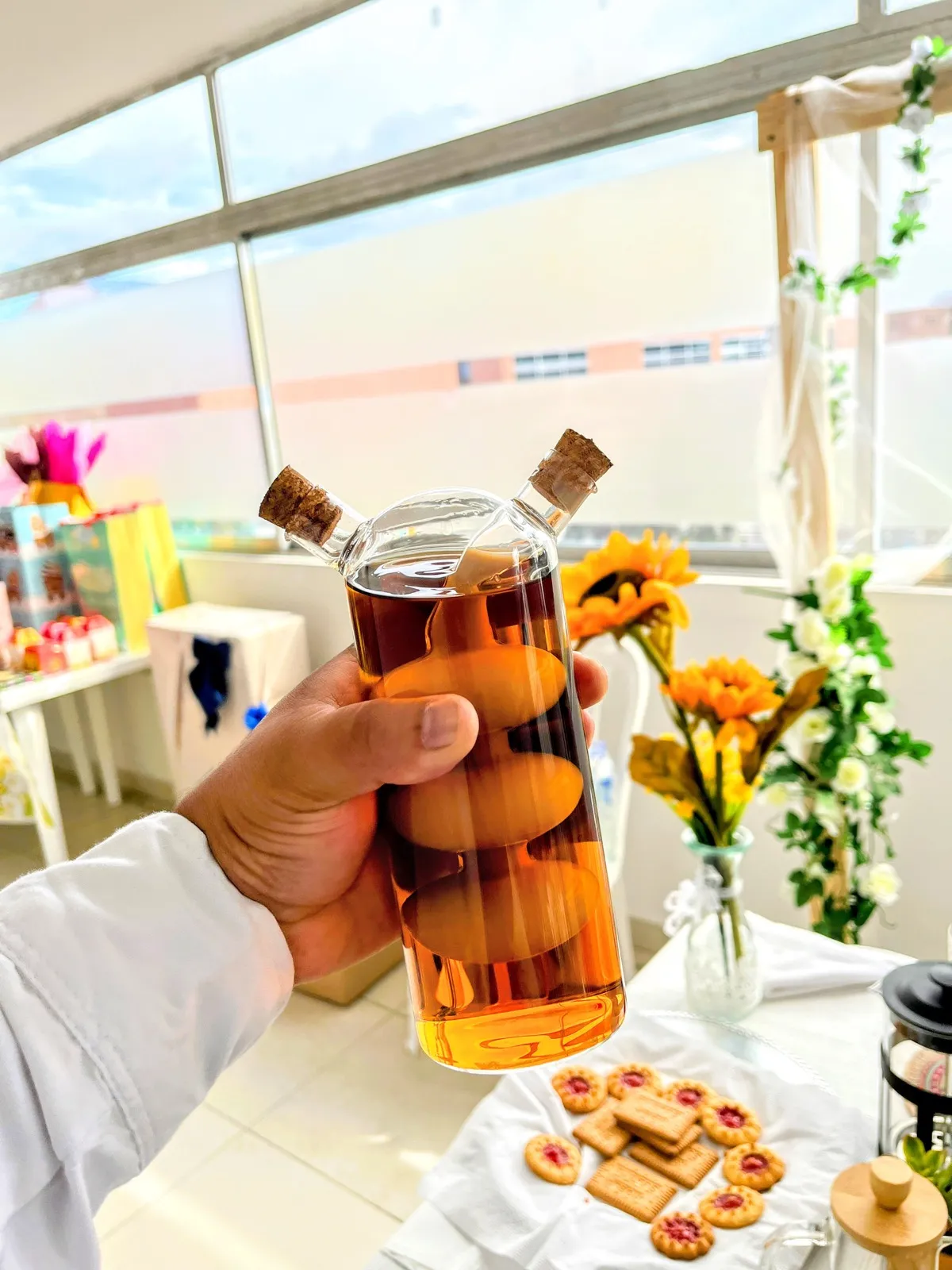 Mano sosteniendo una botella de vidrio con bebida fría junto a galletas y flores amarillas en una mesa de bebidas