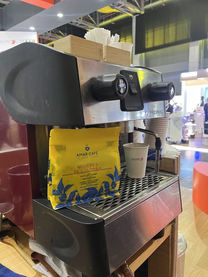 Bolsa de café Amar Café Mujeres de Colombia apoyada en una máquina de espresso dentro de una estación de café