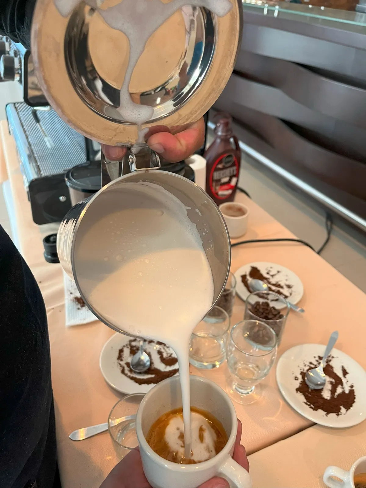 Barista vertiendo leche vaporizada sobre un espresso junto a platos con café molido y vasos para cata