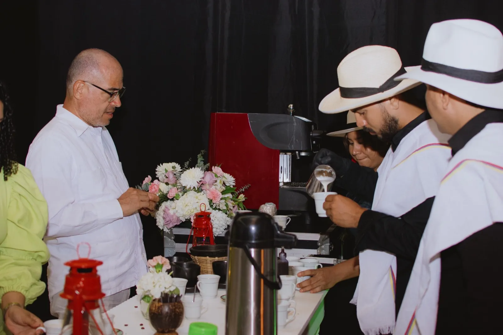 Baristas in traditional Colombian hats and white ponchos serving coffee to guests at a table with flowers, red lanterns and an espresso machine