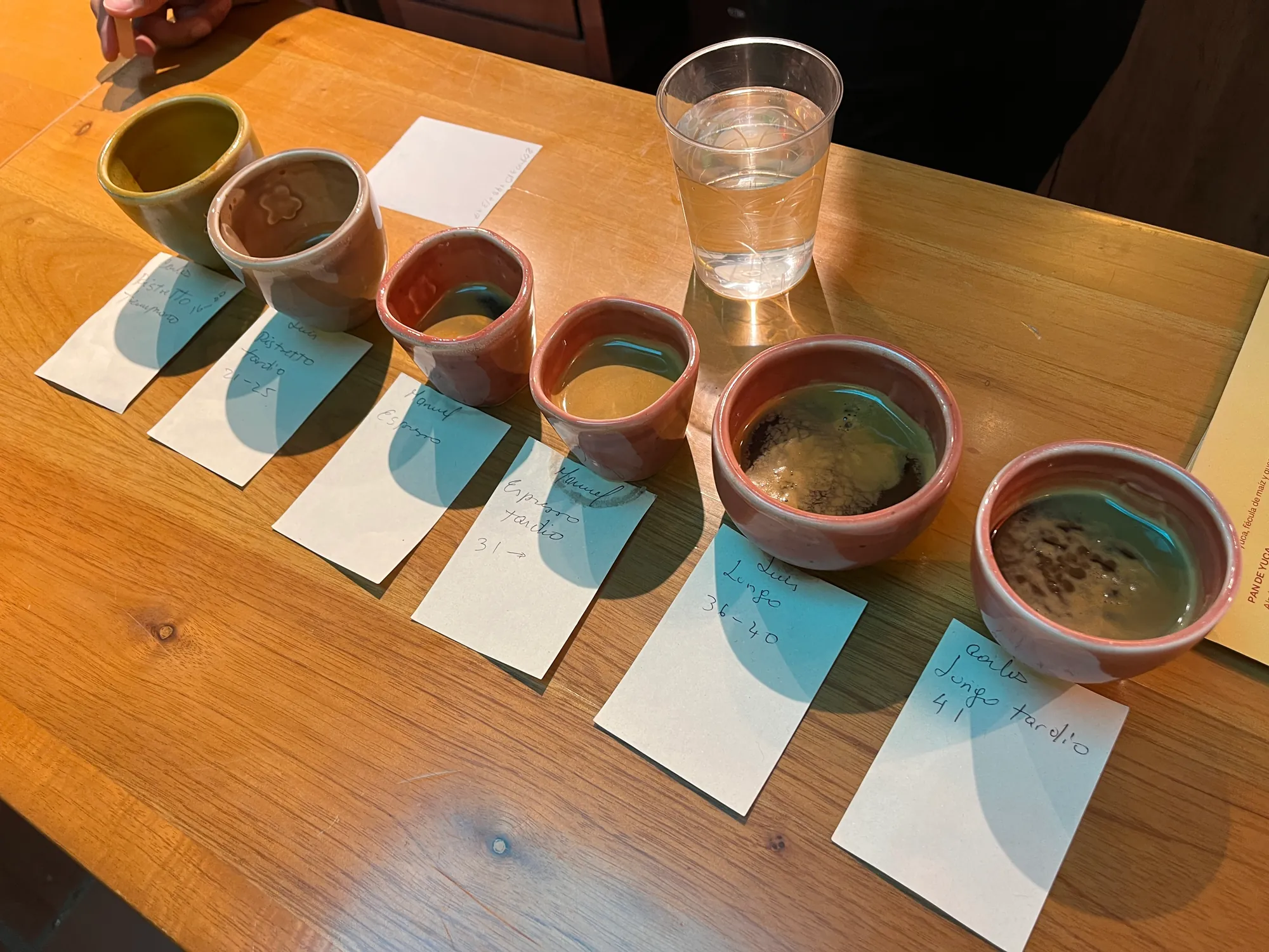 Six small cups of coffee lined up on a wooden table, each with a handwritten tasting card in front of it