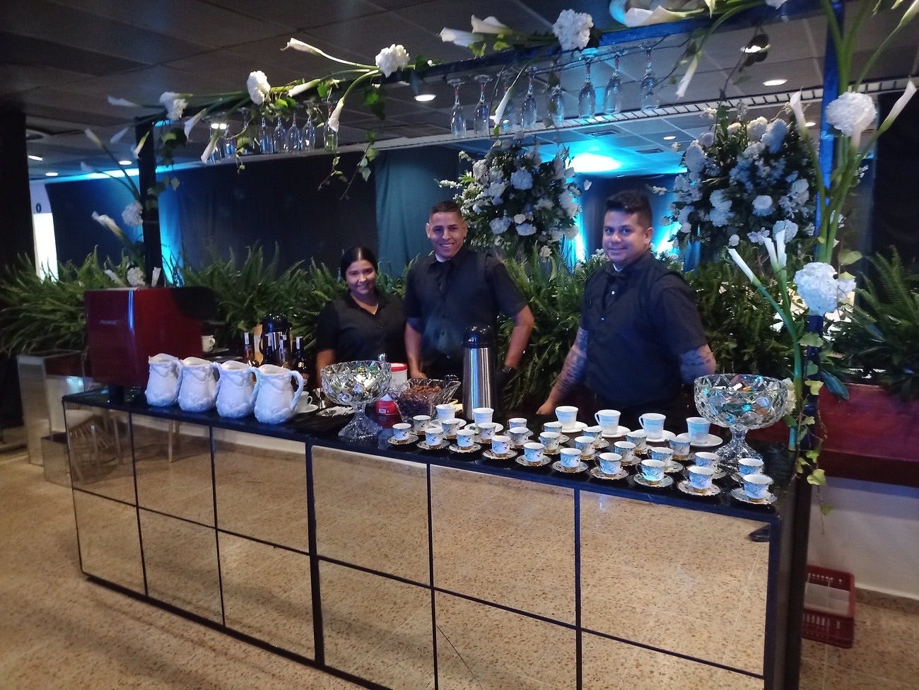 Barra de bebidas con jarras, tazas y copas atendida por tres personas en un evento social
