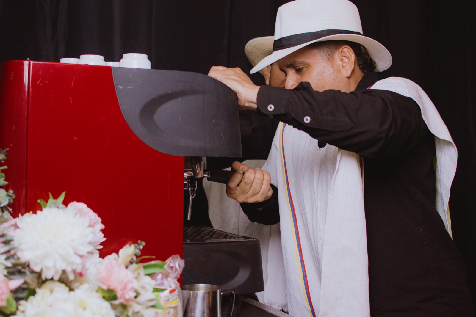 Barista de Experiencia VINTO preparando café en una máquina de espresso roja junto a un arreglo de flores blancas