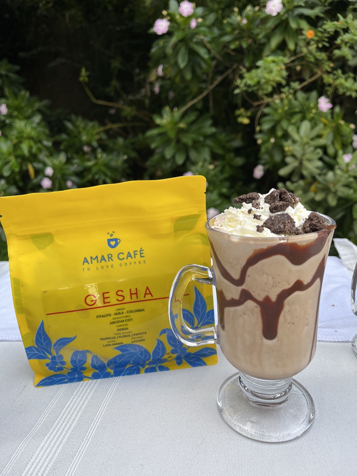 Café frío con crema y chocolate junto a un empaque de Amar Café Gesha en una mesa al aire libre