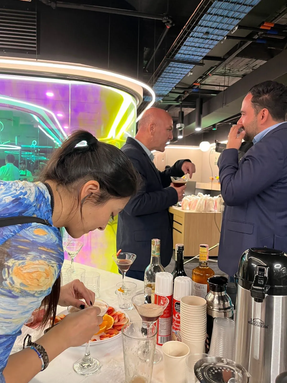 Bartender decorando cócteles de café con rodajas de naranja mientras dos invitados conversan en una oficina